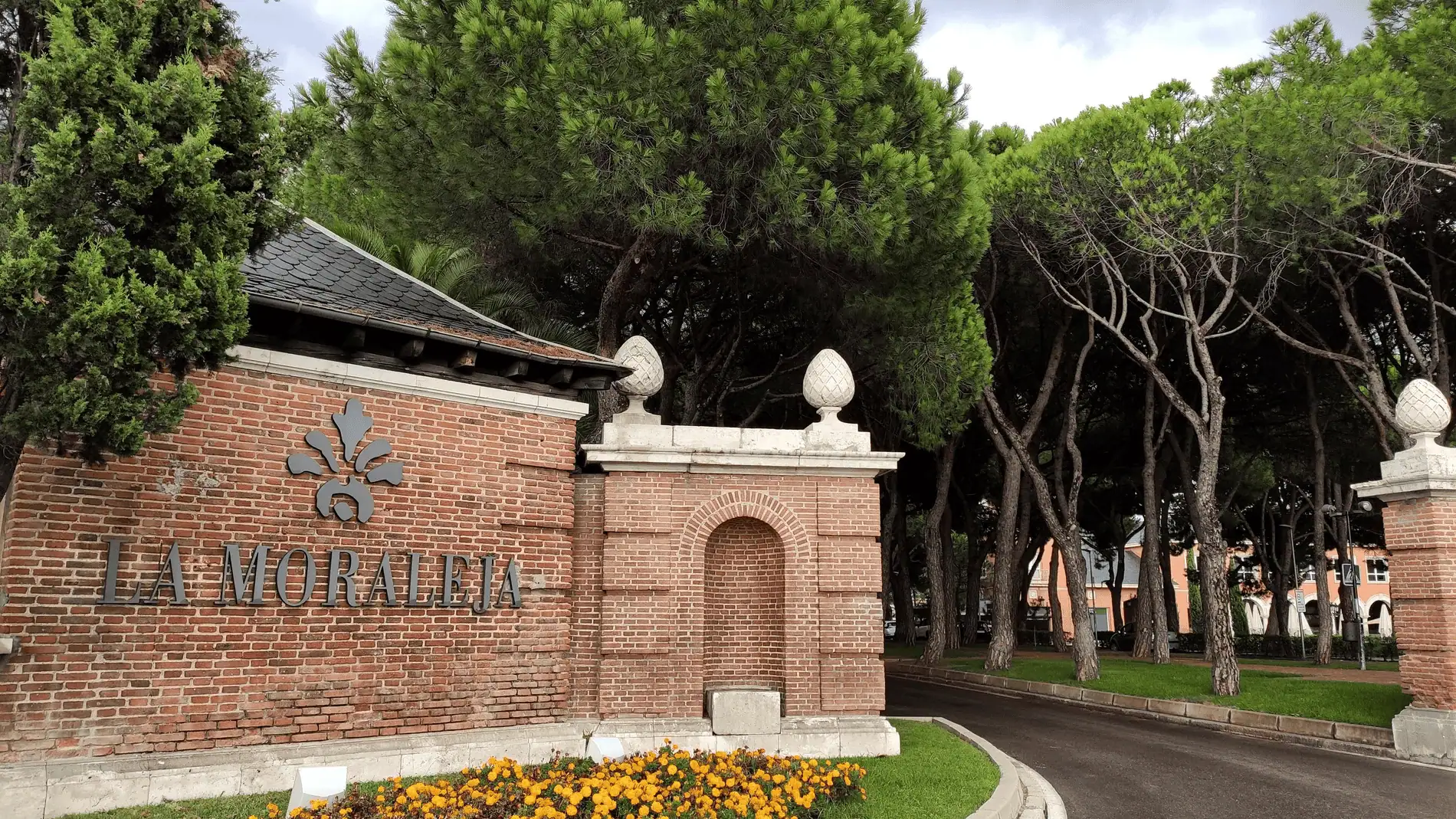 Main entrance to La Moraleja, Madrid’s prestigious residential neighborhood, featuring classic brick architecture and lush pine trees.